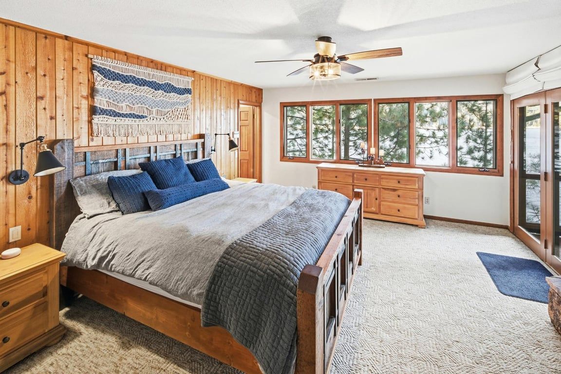 A bedroom with a wooden headboard, a bed with blue and gray bedding, a desk, and a large window with a view of trees.