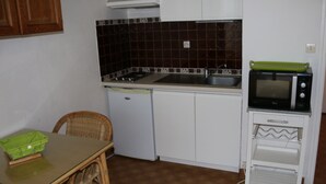 A compact kitchen with a sink, stove, and microwave. There's a wicker chair and a small table with a green bag on it. - Cozy studio with parking in Cap d'Agde, near the sea (Cap d'Agde naturisme)