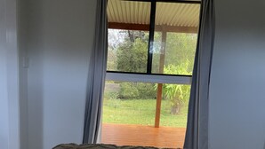 A room with a bed, a window with curtains, and a view of a garden. - Country cabin (Landsborough)