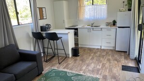 A compact kitchen with a microwave, kettle, and sink. There are two bar stools and a small trash bin. The room has a wooden floor and a green patterned rug. - Country cabin (Landsborough)