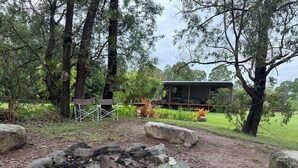 A rustic outdoor area with a fire pit surrounded by rocks, a wooden deck, and a cabin in the background. - Country cabin (Landsborough)