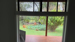 A window with a view of a garden, a wooden deck, and a chair. - Country cabin (Landsborough)