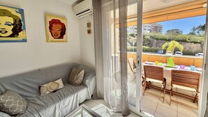 Ein modernes Wohnzimmer mit einem grauen Sofa, zwei Wandbildern und einer Glastür, die auf einen Balkon mit Tisch und Stühlen führt.