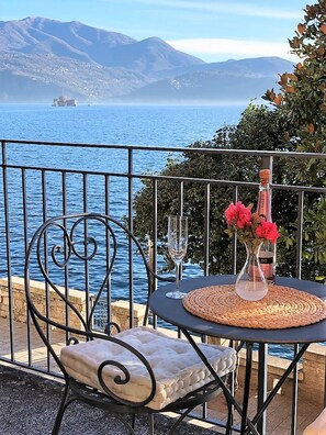 A balcony with a view of the sea and mountains, a table with a vase of flowers, a wine glass, and a chair.