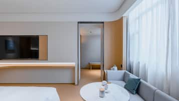 A modern hotel room with a bed, a sofa, a dining table, and a TV mounted on the wall.