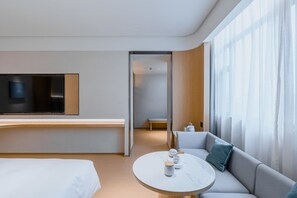 A modern hotel room with a bed, a sofa, a dining table, and a TV mounted on the wall.