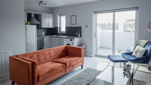 Un apartamento moderno con una sala de estar que incluye un sofá, una silla y una mesa de centro de vidrio. Hay una cocina con armarios blancos y un ventanal con vistas a la ciudad.