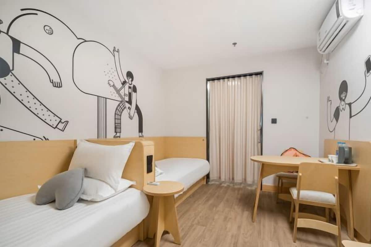 A hotel room with two single beds, a small table with chairs, an air conditioning unit, and a window with curtains.