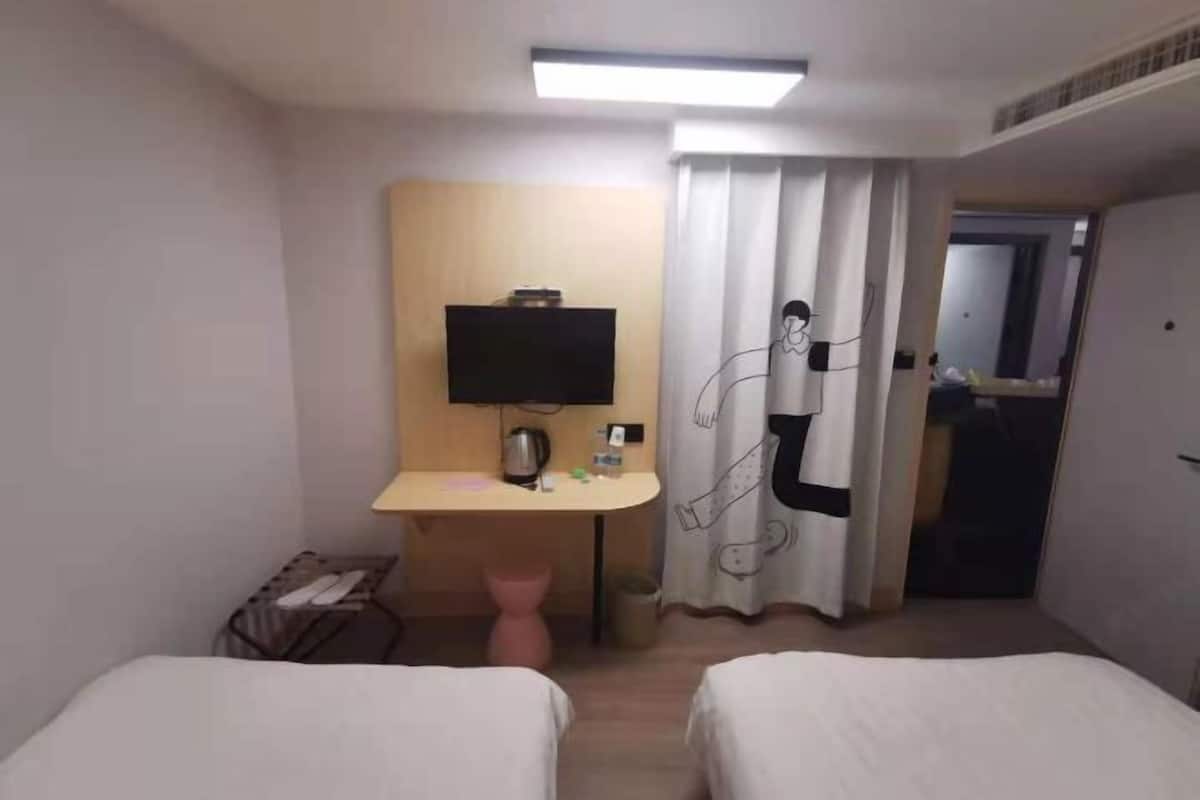 A hotel room with two beds, a desk with a flat-screen TV, a pink chair, and a curtain with a cartoon character design.