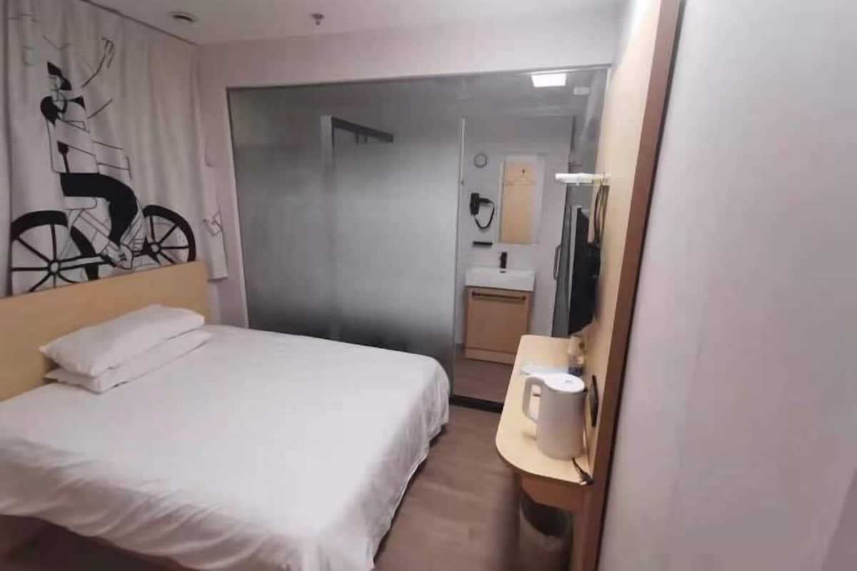 A hotel room with a bed, a small table with a coffee maker, a mirror, and a bathroom visible through a glass door.