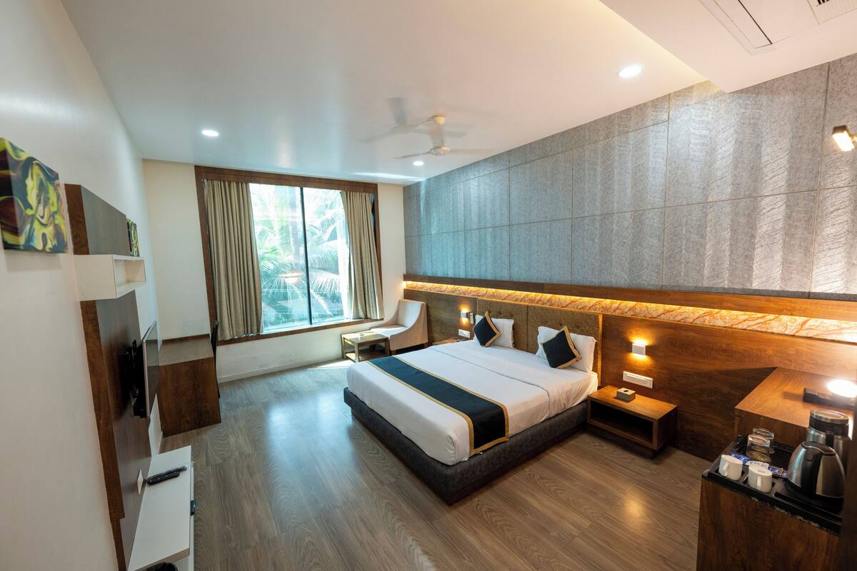 Quarto de hotel moderno com cama, mesas de cabeceira, escrivaninha, televisão e janela com cortinas.