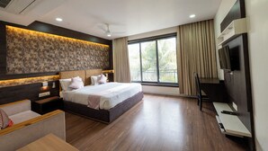 A modern hotel room with a large bed, a TV, a desk, and a window with curtains. - The Grand Astoria Somnath (Veraval)