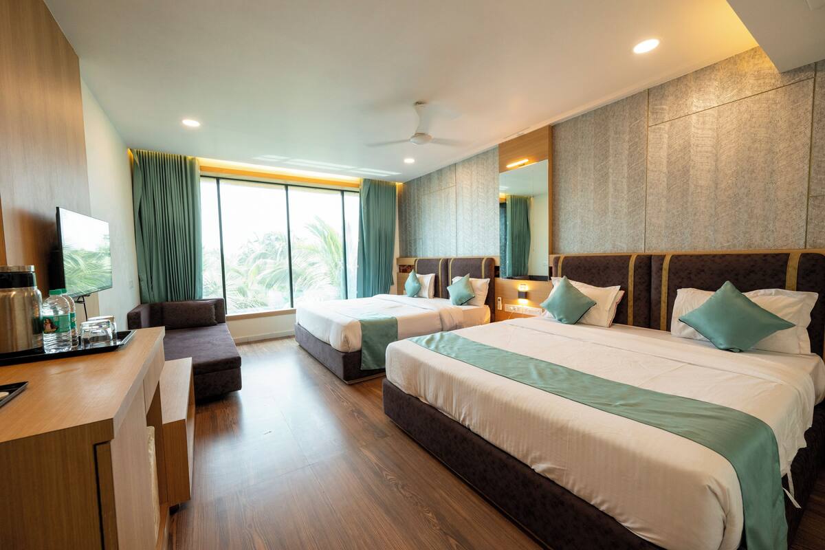 Quarto de hotel com duas camas, uma escrivaninha, um sofá, uma televisão e uma janela grande com cortinas.