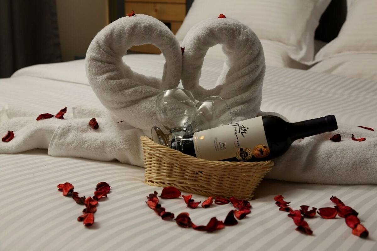 Una cama con ropa blanca, una cesta de mimbre con una botella y copas, y pétalos de rosa roja esparcidos sobre la cama.