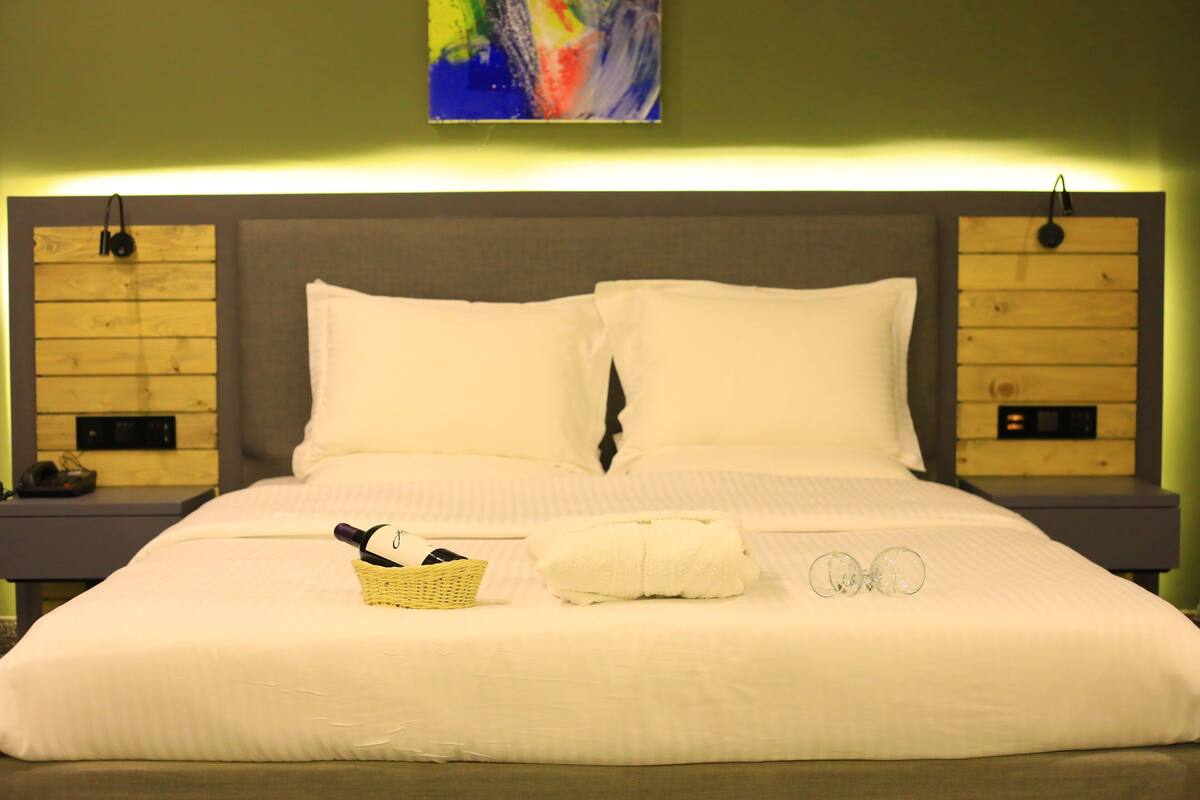 Una cama bien hecha con sábanas blancas, una mesita de noche con lámpara, y un cuadro abstracto de colores en la pared.