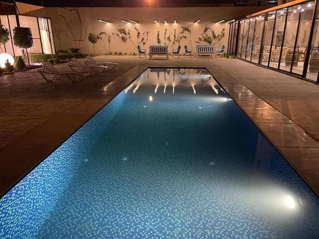 Une piscine entourée de chaises longues et d’un espace salon, le tout bordé d’une terrasse bien éclairée, agrémentée de plantes et dotée d’une architecture moderne.