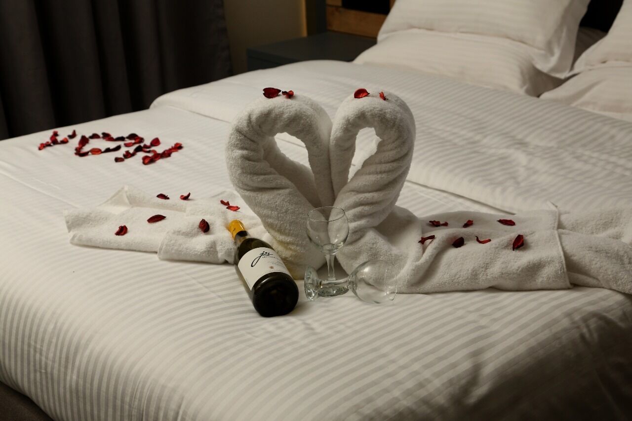 A bed with a heart-shaped towel arrangement, a bottle of wine, and empty wine glasses on a white bedspread.