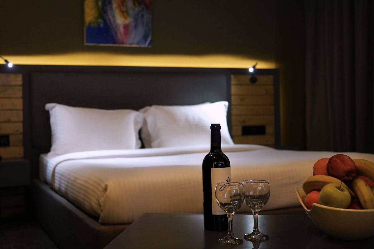Habitación de hotel con cama, mesita de noche con una botella de vino, dos copas y un frutero.