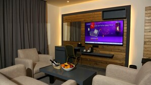 A modern living room with a flat-screen TV mounted on a wooden wall, a coffee table with fruits and a bottle, and comfortable seating. - Aitch Boutique Hotel - an LIH Hotel (Madaba)