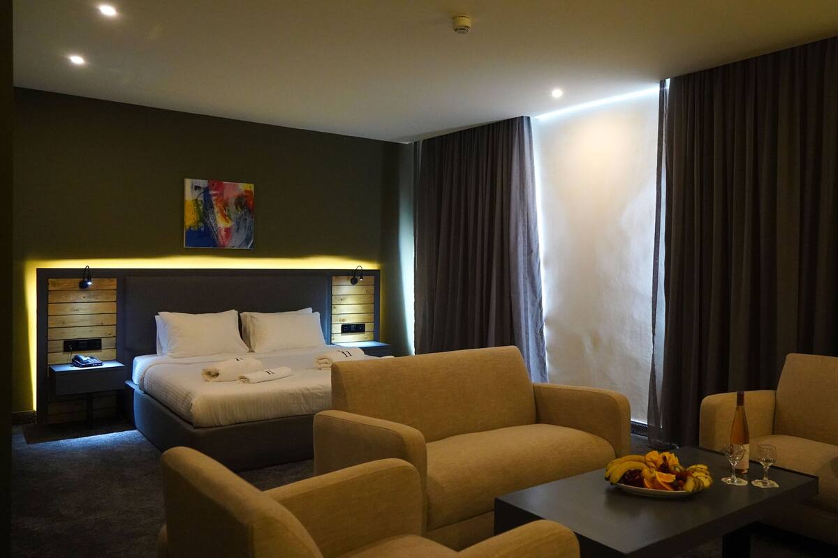 Una habitación de hotel con cama, dos sillones, una mesita de centro con fruta y bebidas, un cuadro en la pared y una mesita de noche con lámpara.