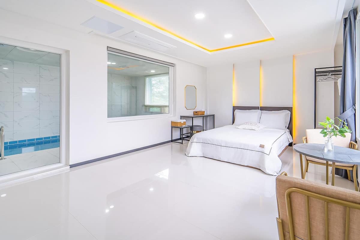Une chambre moderne avec un grand lit, une table de chevet, une chaise, une petite table avec une plante et une grande fenêtre.