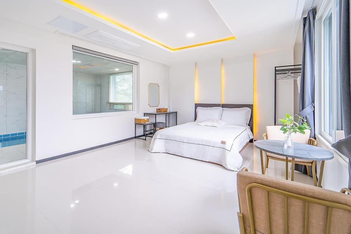 Une chambre moderne avec un grand lit, une table de chevet, une chaise, une petite table avec une plante et une grande fenêtre.