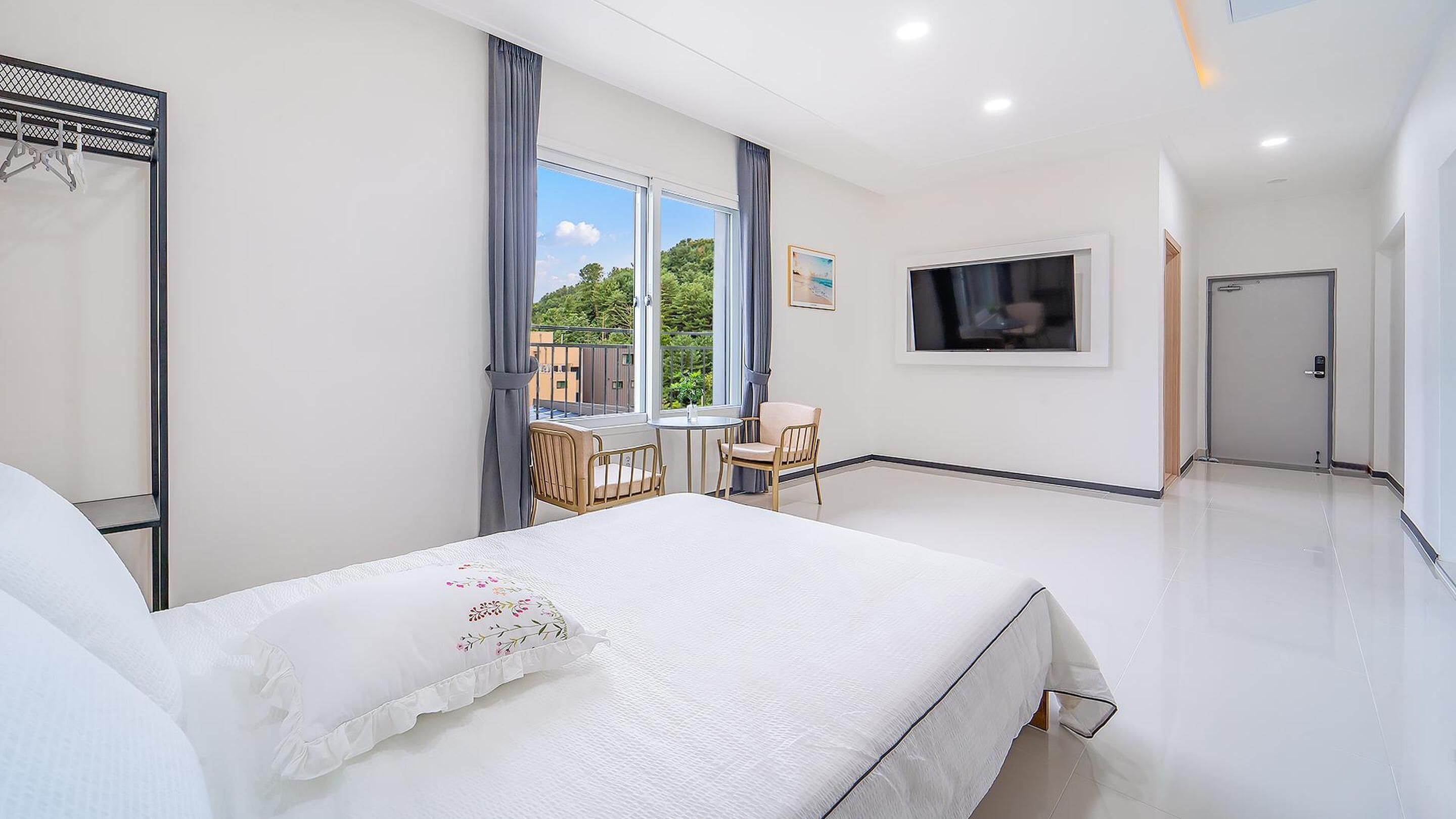 A hotel room with a bed, a small table with chairs, a TV mounted on the wall, and a balcony with a view of greenery.
