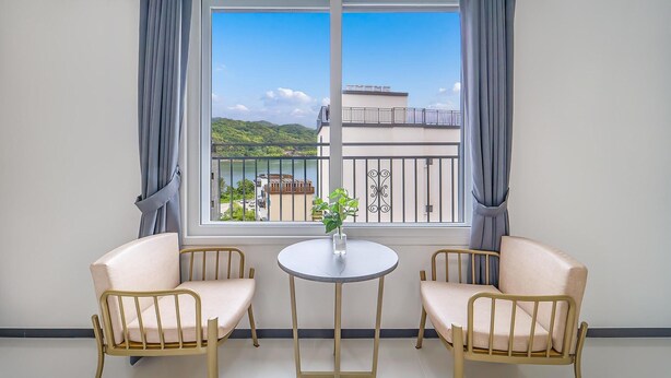 Een kamer met twee beige stoelen, een kleine ronde tafel en uitzicht op een balkon met een hekwerk en beplanting.