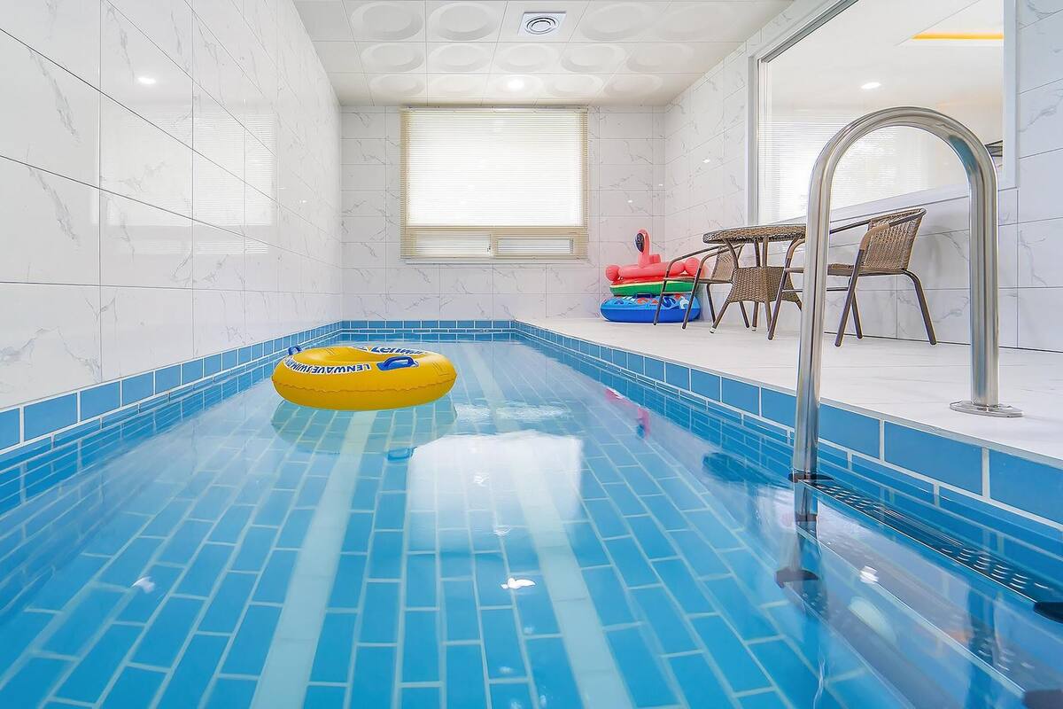 Une piscine avec un sol carrelé de carreaux bleus, un flotteur jaune et, au fond, une fenêtre avec des stores.