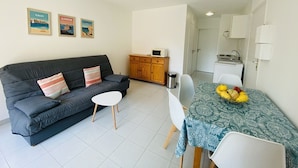 A compact living space with a dark grey sofa, a small white table, and a dining table with a patterned tablecloth. There are three framed pictures on the wall, a wooden cabinet, and a kitchenette area. - Cozy 1-Bedroom Apartment with Terrace, 100m from Beach, Parking Included - Calvi (Calvi)