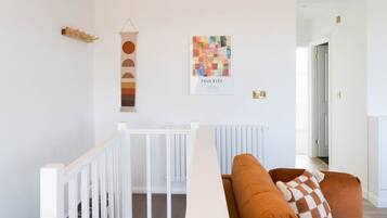 Een kamer met een wit kinderbed, een bruine bank met een geruit plaid, en een wand met een kleurrijk abstract schilderij en een houten decoratie.