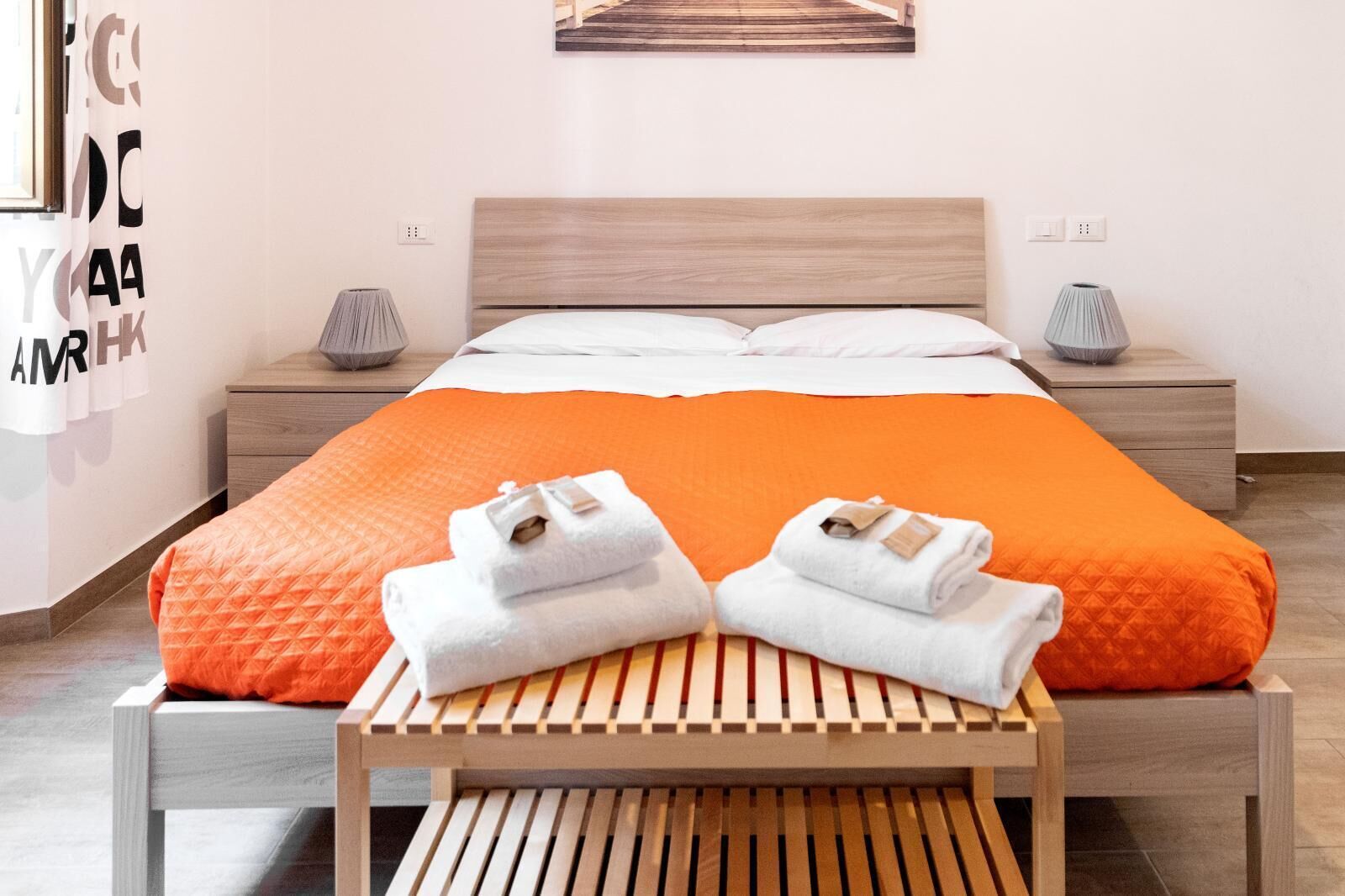 A neatly made bed with an orange blanket, two white towels on a wooden bench, and a wooden headboard.