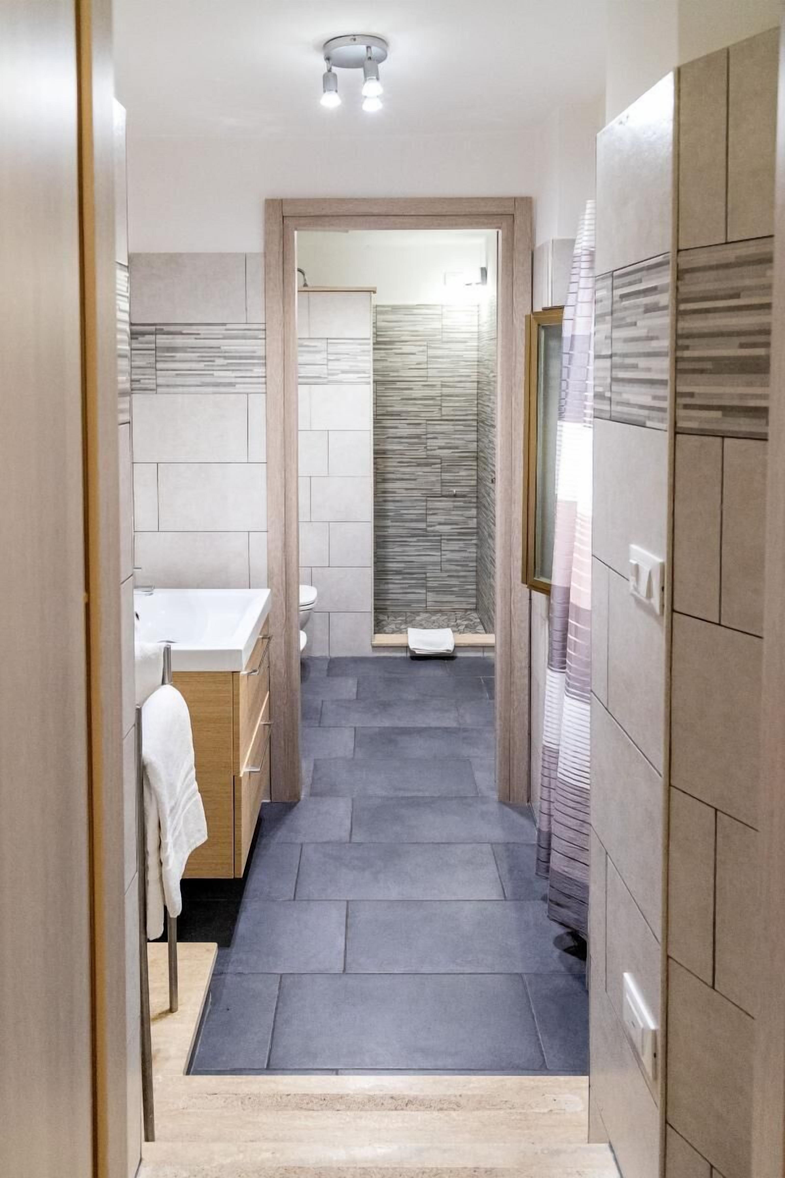 A narrow bathroom with a white sink, a toilet, and a shower area.