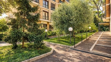 A residential building with a brick facade, multiple balconies, and a well-maintained garden area with trees and a paved walkway.