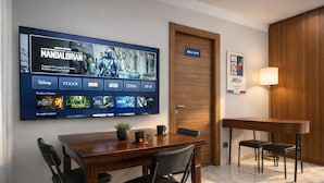 A hotel room with a large flat-screen TV displaying a movie, a wooden door labeled 'Blue room,' a small table with chairs, and a lamp. - AR ROOMS ROME   blue room superior (ROMA)