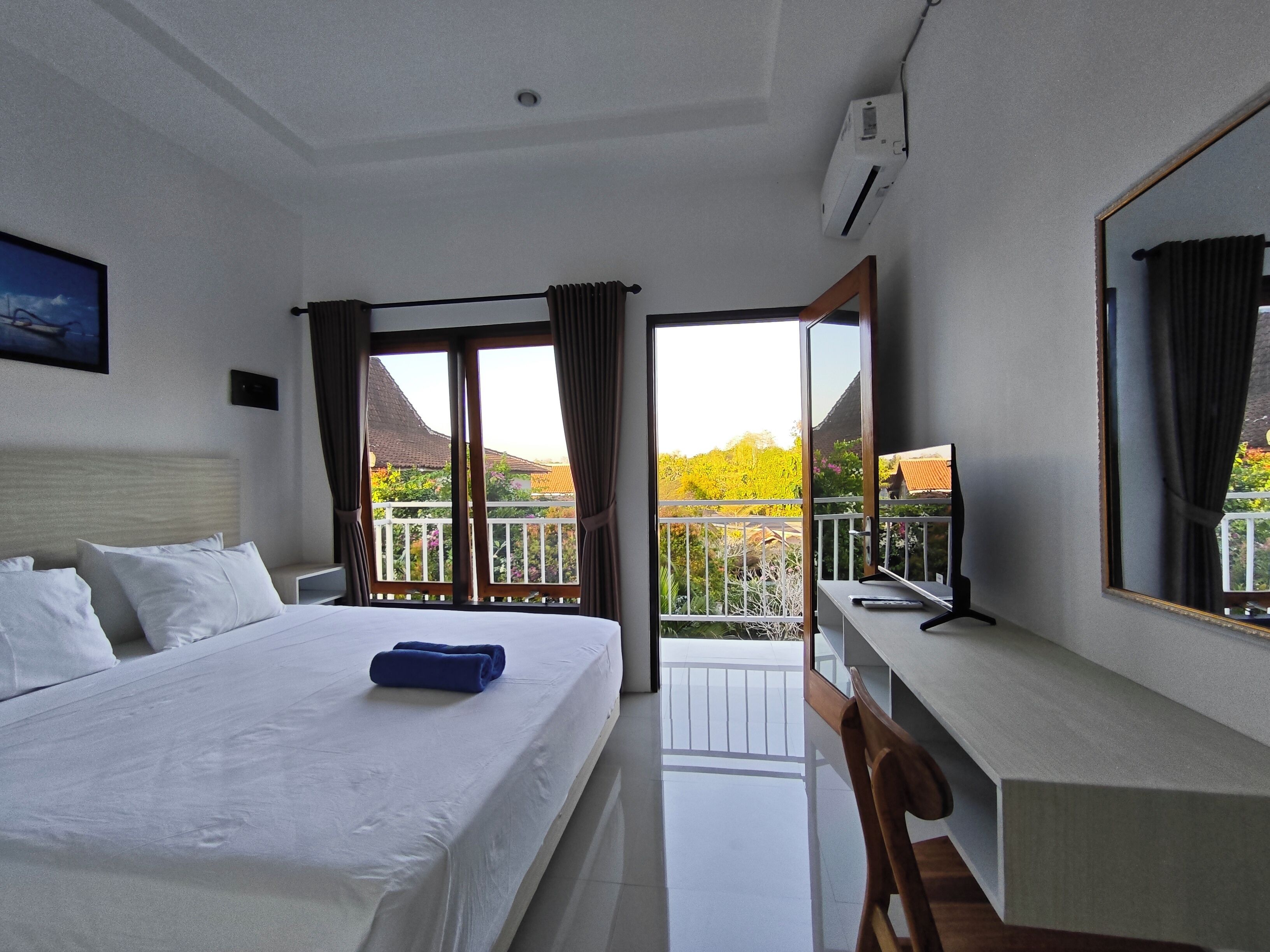Habitación de hotel con cama, escritorio, silla, un espejo grande y balcón con vistas a zonas verdes y edificios.