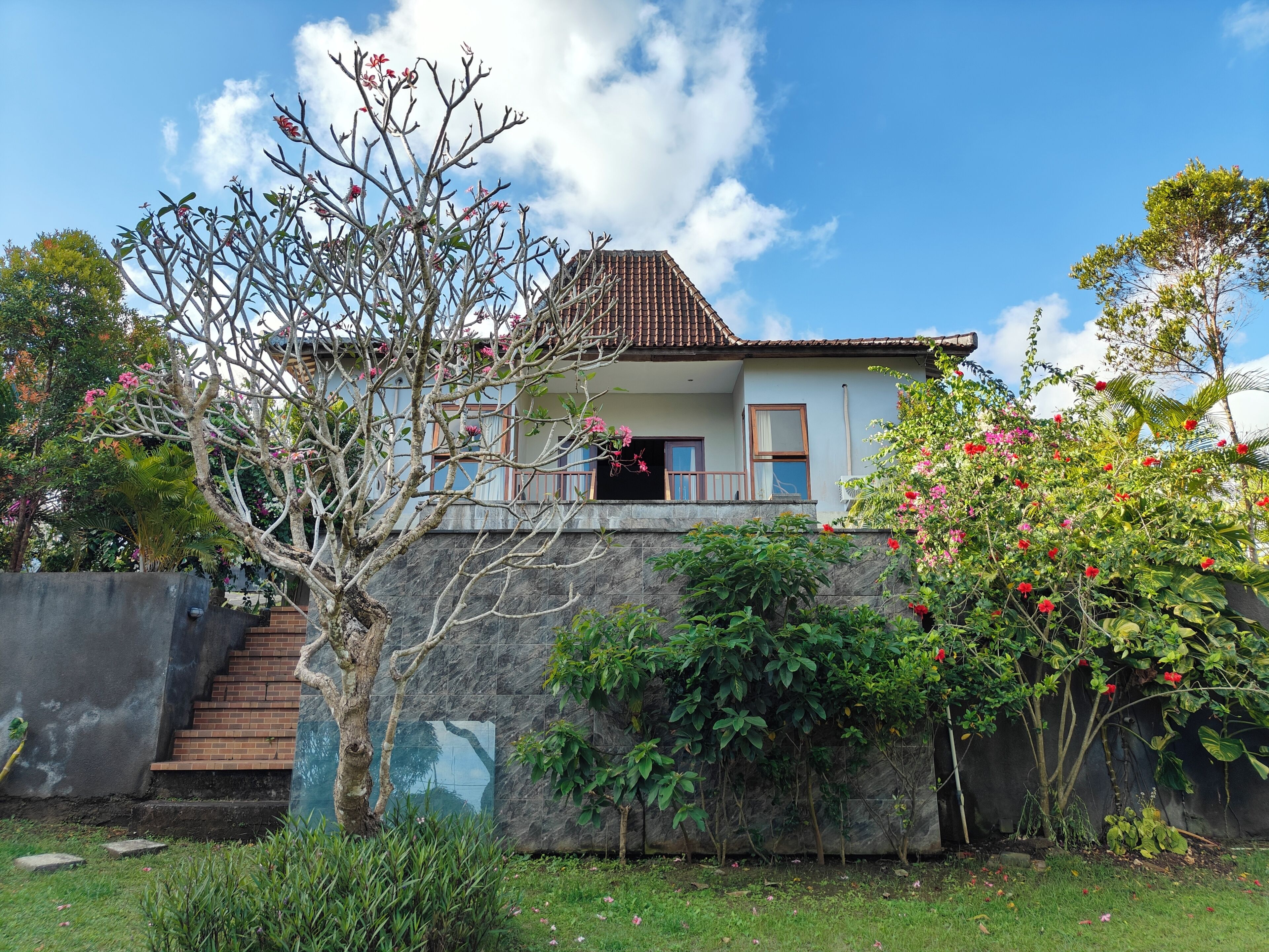 A house with a tiled roof, a staircase leading to a balcony, and lush greenery around.