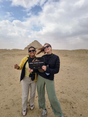 Dos personas sosteniendo un cartel con el texto 'JANA TOURS EGYPT', de pie en un desierto con dunas de arena al fondo.
