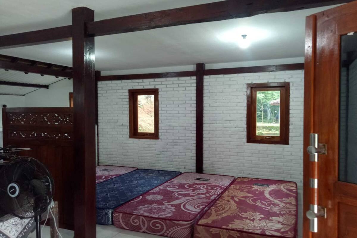A room with a white brick wall, two windows, and a wooden door. There are three mattresses with floral patterns on the floor.
