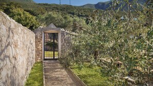 A stone pathway leading to a gate, surrounded by olive trees and a mountainous backdrop.
