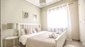 A bedroom with a bed, a bedside table, a lamp, and a window with curtains. - Grand Meteora Central Luxury Apartment (Kalabaka)