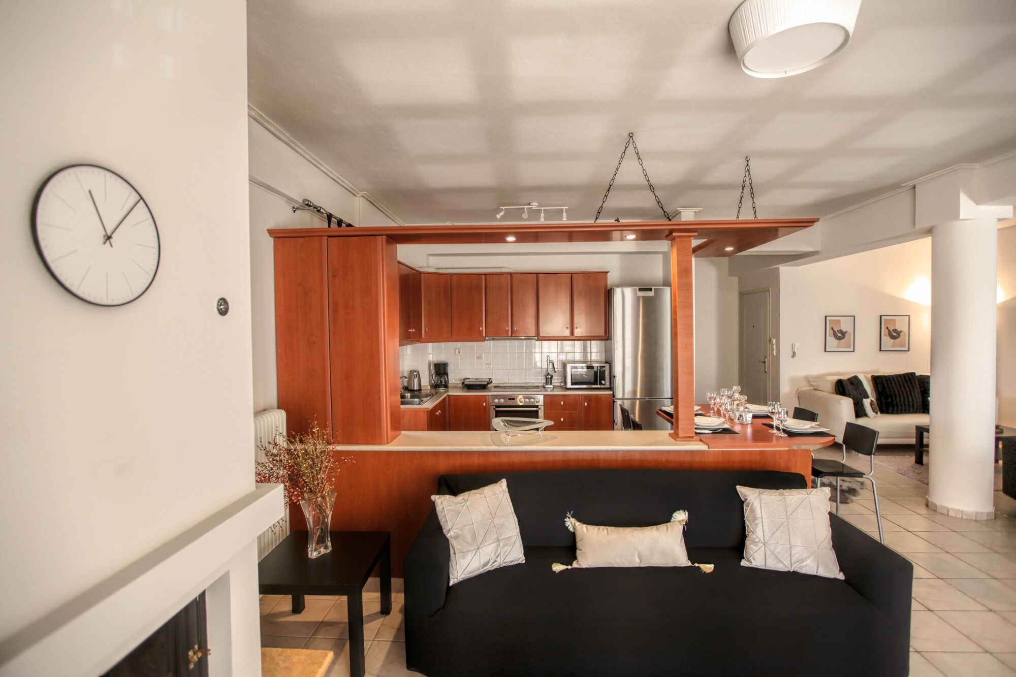 A modern living room with a black sofa, a dining table set for a meal, and a kitchen area with wooden cabinets and stainless steel appliances.