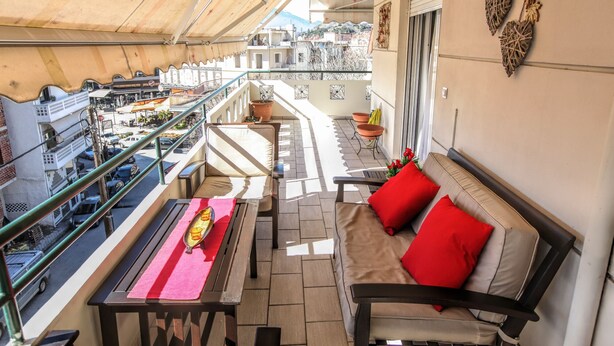 A balcony with a sofa and table, a view of the city, and a railing. - Grand Meteora Central Luxury Apartment (Kalabaka)