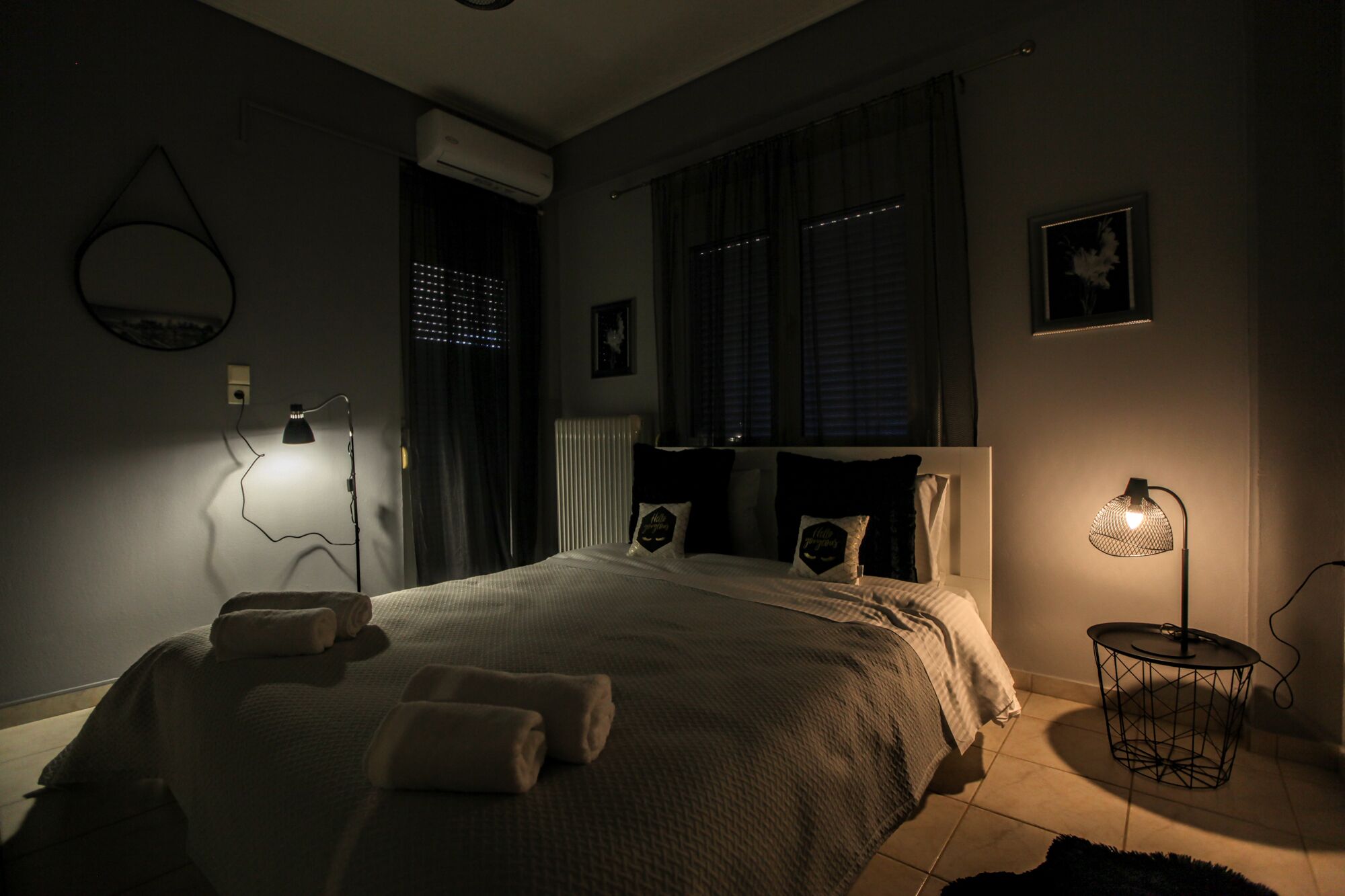 Quarto com pouca luz, com uma cama, duas luminárias de apoio, um radiador e uma mesinha com abajur.