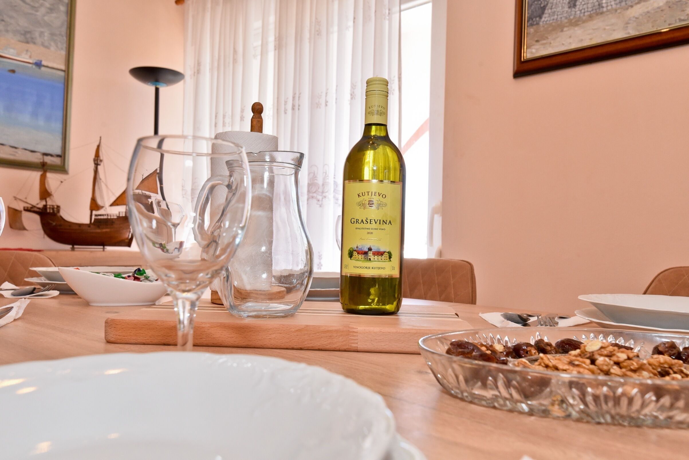 A dining table with a bottle of Kutjevo Graševina wine, two wine glasses, a bowl of granola, and a ship model.