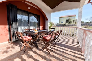A covered outdoor dining area with a table and chairs, a view of a building in the background, and a tiled floor.