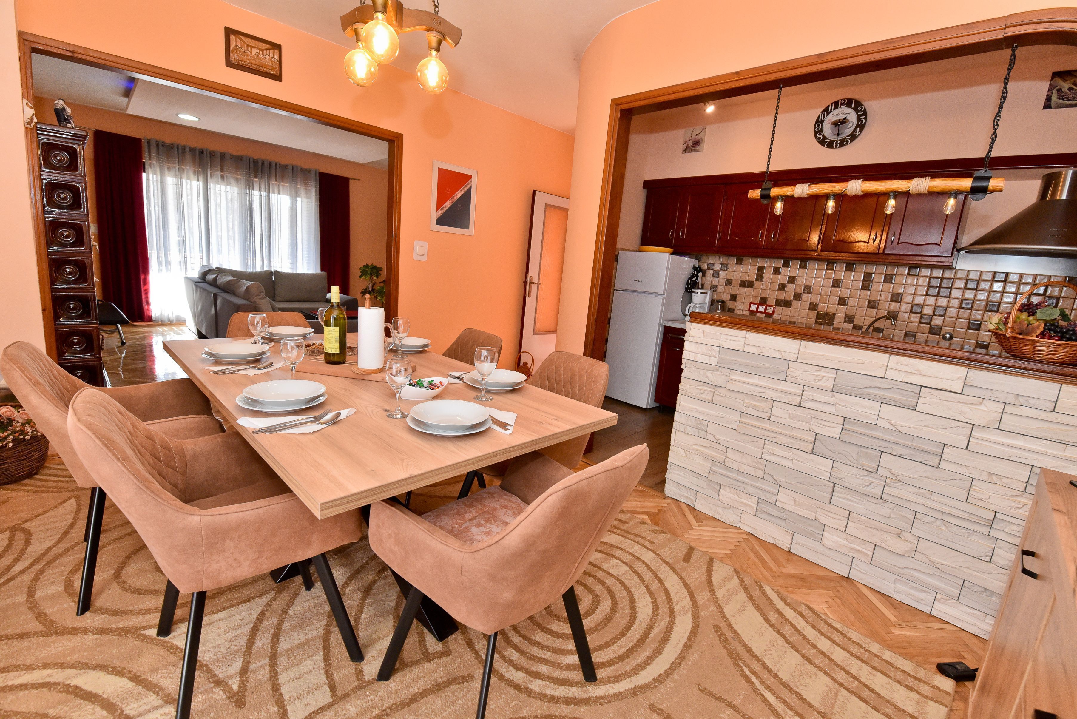 A dining area with a wooden table set for four, surrounded by cushioned chairs. A kitchen with a stone backsplash and wooden cabinets is visible in the background.