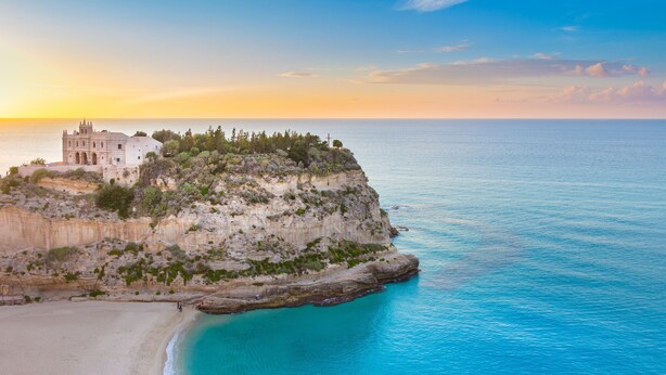 A coastal building with a beach in the foreground and a cliff with vegetation. - VOI Le Muse Essentia (Zambrone)