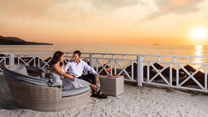 A couple seated on wicker chairs by a beach with a sunset view, white railings, and a drink on a table. - VOI Le Muse Essentia (Zambrone)