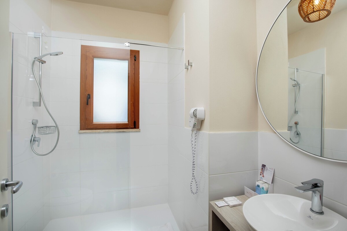 A modern bathroom with a glass shower enclosure, a wooden-framed window, a white sink, and a large mirror.